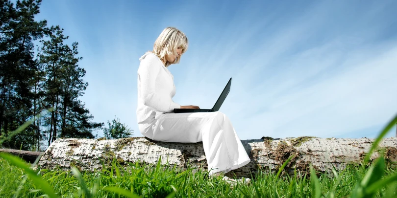 vrouw zit op boomstam in de buitenlucht te werken op haar laptop