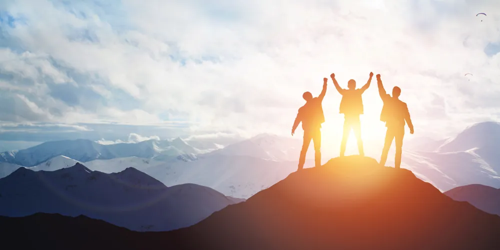 3 mensen bovenaan op een rots met zonsondergang en handen in de lucht, leiderschap